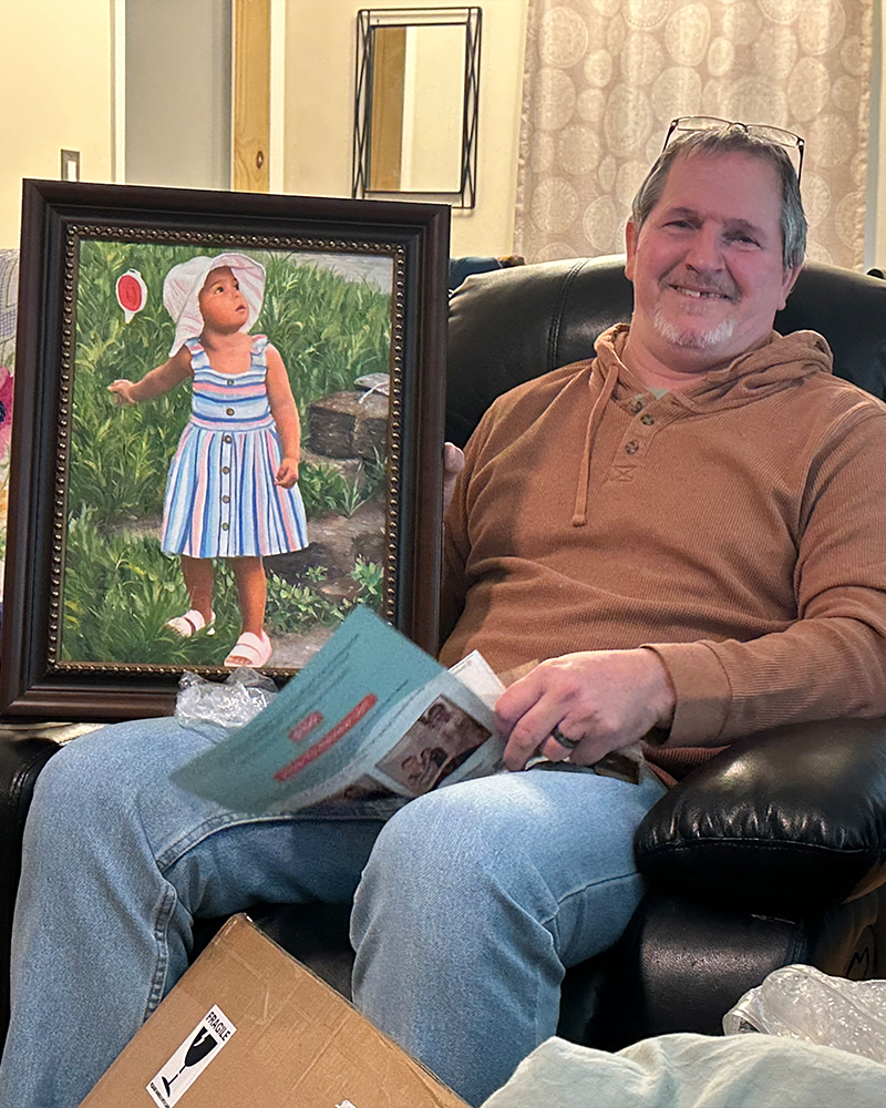 Man seated beside framed custom portrait of a young girl in a garden